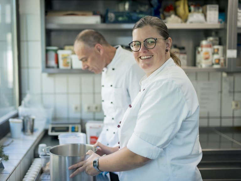 Eine Frau lächelt in die Kamera, während sie mit einem Mann in der Küche arbeitet.