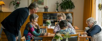 Haus Schillerstraße Eine Mitarbeiterin unterhält sich mit einer Gruppe Senioren, die am Esstisch sitzt.